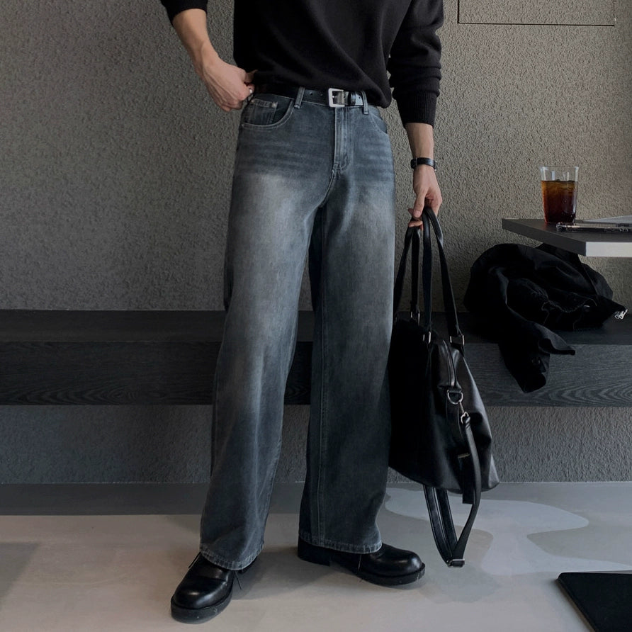 Person wearing gray jeans and black shoes holding a black bag in an indoor setting.